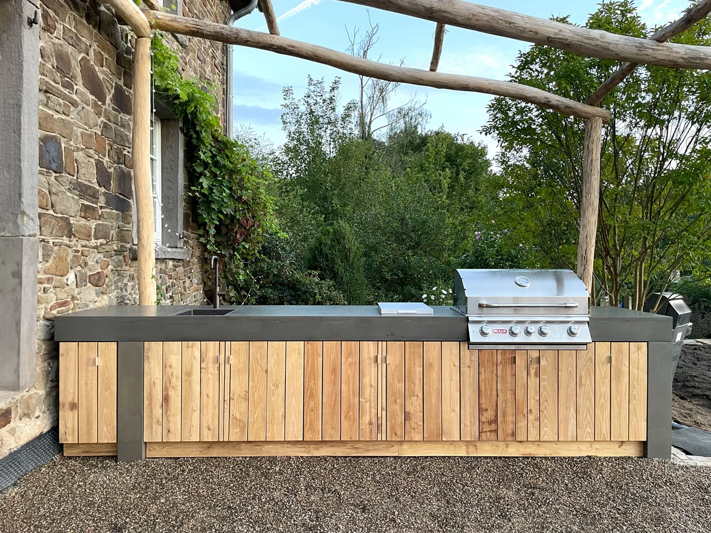 Außenküche mit Beton-Arbeitsplatte, Holzfronten, Spüle und Gasgrill vor einer Natursteinwand
